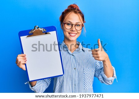 Young redhead woman holding clipboard with blank space smiling happy and positive, thumb up doing excellent and approval sign 