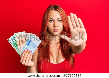 Young beautiful redhead woman holding south korean won banknotes with open hand doing stop sign with serious and confident expression, defense gesture 