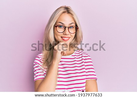 Beautiful caucasian blonde girl wearing casual clothes and glasses smiling looking confident at the camera with crossed arms and hand on chin. thinking positive. 