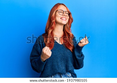 Young beautiful redhead woman wearing casual sweater and glasses over blue background very happy and excited doing winner gesture with arms raised, smiling and screaming for success. celebration Royalty-Free Stock Photo #1937980105