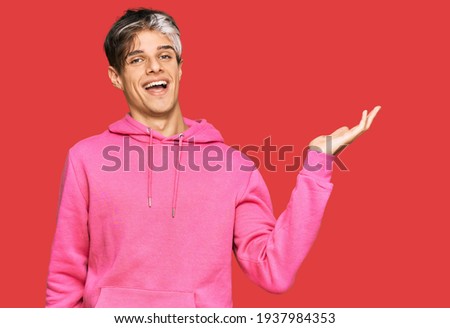 Young hispanic man wearing casual pink sweatshirt smiling cheerful presenting and pointing with palm of hand looking at the camera. 