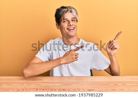 Young hispanic man wearing casual clothes sitting on the table smiling and looking at the camera pointing with two hands and fingers to the side. 