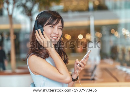 Asian woman using smart mobile phone and listening the music via Head cover headphones in department store or shop store, entertainment and lifestyle concept Royalty-Free Stock Photo #1937993455