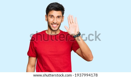 Young handsome man wearing casual red tshirt waiving saying hello happy and smiling, friendly welcome gesture  Royalty-Free Stock Photo #1937994178