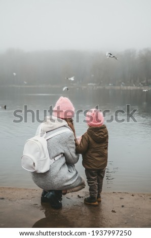 Mom in grey coat and daughter in green jacket both with pink caps are feeding the ducks swimming on the lake in city park