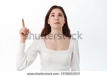 Sad girl looking up with regret and disappointment, pointing finger aside on top banner, longing for something, wanting to have item for herself, standing against white background