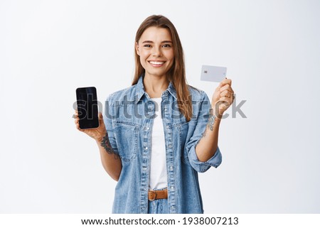 Online shopping. Portrait of smiling blond girl recommending shop, showing empty smartphone screen and plastic credit card, standing over white background