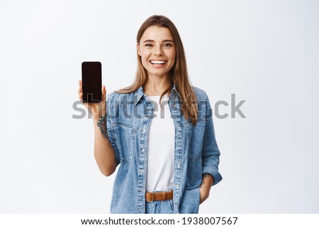 Portrait of attractive young woman with white smile, showing empty smartphone screen, demonstrate an app or online shopping store, studio background