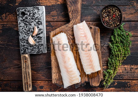 Raw cod loin fillet steak on wooden board with butcher cleaver. Dark wooden background. Top view
