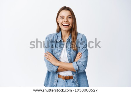 Happy blond girl in casual clothes, laughing from funny joke, looking at camera carefree and having fun, standing with arms crossed on chest, white background
