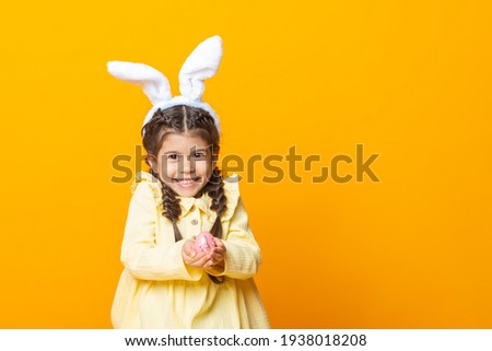 Happy Easter, kids. A cute little girl in the ears of an Easter bunny smiles and holds a pink egg on a yellow background. The concept of Easter.Space for text.  Funny smiling child