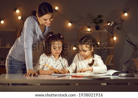 Portrait of happily smiling young caucasian mother watching two adorable twins daughters children drawing picture sitting at desk. Happy family and motherhood, evening home fun activity together