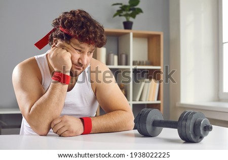Funny sceptic lazy curly man looking thoughtfully at small barbell unsure whether he needs sports exercise. Sad fat man afraid of failure has no motivation to start training with dumbbells at home Royalty-Free Stock Photo #1938022225