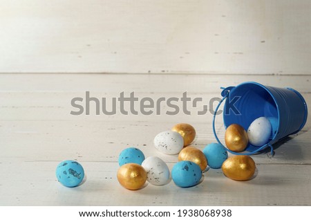 Golden, white and blue Easter eggs with blue bucket on rustic wooden white background. Trendy Easter composition. Greeting card, poster, flyer, promotion, article. Copy space. Minimal Easter concept.