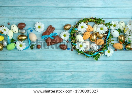 Chocolate Bunny with a satin ribbon surrounded by sweet treats, pieces of eggs, colorful candies. A wreath of twigs. Light blue wooden background and copy space. Modern greeting card.