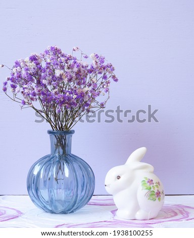 Composition of purple colored baby's breath (gypsophila) in a blue glass vase and a porcelain white rabbit with painted flowers sit atop a table top.