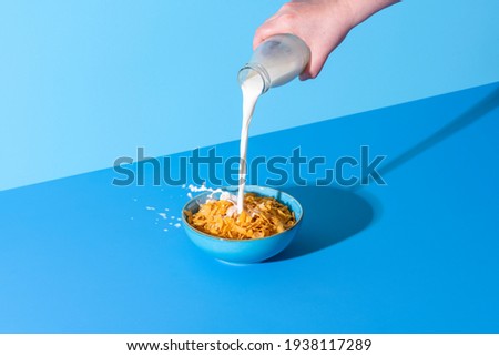 Pouring milk from a bottle into a bowl with cornflakes cereals. Bowl with corn cereals and milk isolated on a blue background Royalty-Free Stock Photo #1938117289