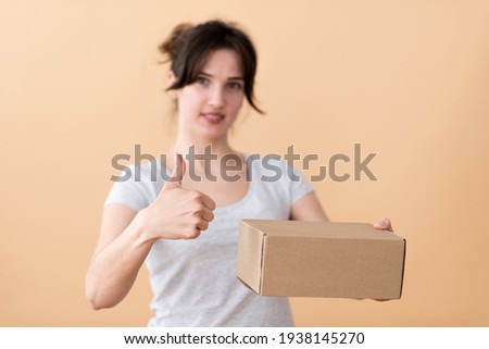 Young european woman advertises cardboard box in the foreground, gestures to the quality with her finger up, advertising concept