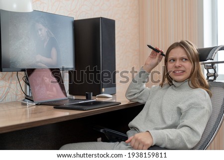 the girl thoughtfully scratches her head with a stylus while sitting at a computer table and processing her photo using a graphics tablet