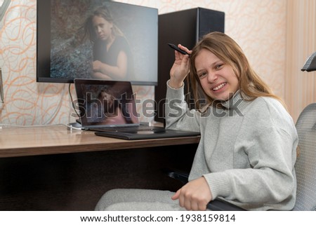 an emotional girl processes her photo while sitting at a table on which there is a graphics tablet, a laptop and a large monitor