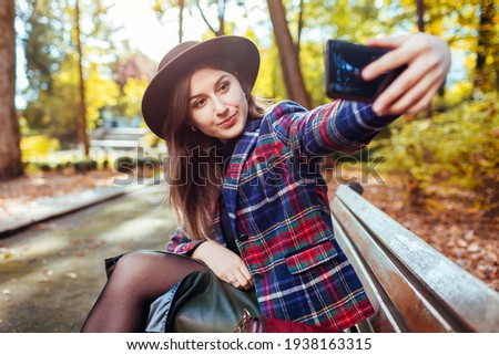 Stylish woman in hat relaxing in spring park. Girl taking selfie on smartphone sitting on bench outdoors.