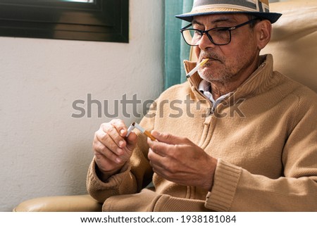 Elderly man wearing a hat and black glasses, holding in his mouth a plastic cigarette to stop smoking, and in his hands a cigar that he has broken who looks thoughtful as a sign of rejection of tobacc