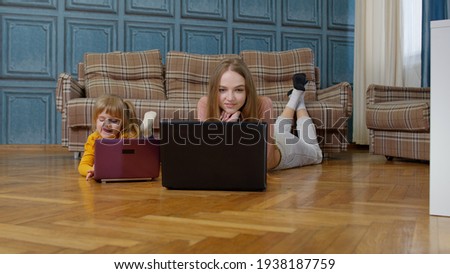 Young mother and small preschool daughter child sit on warm wooden floor in living room, parent focused on working typing on laptop. Girl kid watching lessons online, cartoons movies, studying at home