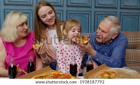 Multigenerational family having lunch party, feeding child girl with pizza, laughing, enjoying meal together at home. Grandparents with young woman and kid. Celebration holidays, weekends, birthday