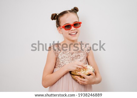 A teen girl with glasses to watch funny 3D movies eats popcorn stands against a white wall. Home or online cinema. harmful snacking.