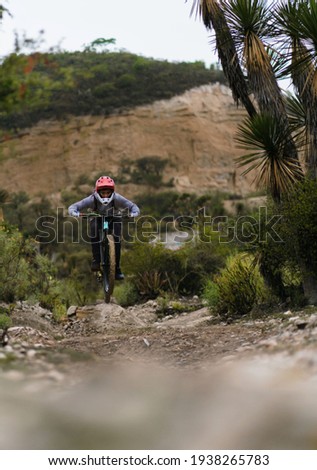 Young mexican practicing mountain bike jumping in the hills of Mexico. He wears sports clothes and red helmet, there is a large mountain in the background