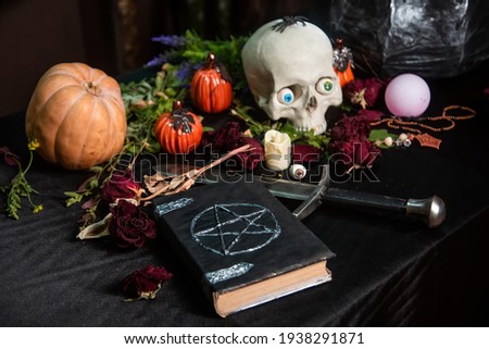 Focus on a black book with a pentagram lying on a black table against a background of a skull and pumpkin. Halloween magic ritual concept.