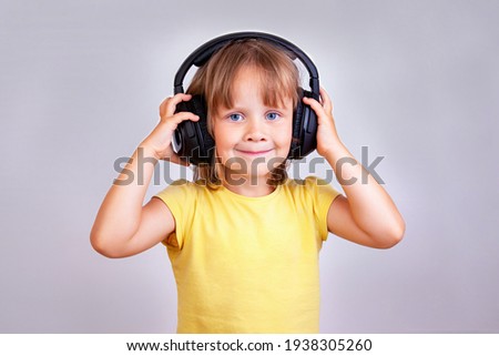 A little cute girl in a yellow T-shirt with large headphones on her head stands on a gray background.