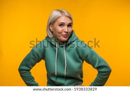 Photo of a bright girl in a dark green hooded jacket which stands against the backdrop of a bright yellow-orange wall and smiles funny on camera. High quality photo
