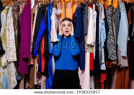 Perplexed beautiful Caucasian teenage girl in clothing store. he doesn't know what to choose. looking at camera. Beautiful young woman near rack near hangers with clothing. 