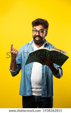Young Indian happy man wearing eyeglasses reading book and smiling isolated over yellow background