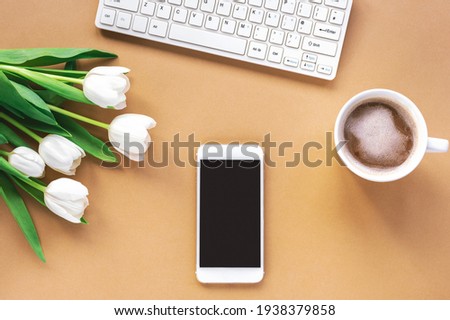 Workplace with coffee cup, phone, computer keyboard and white tulips. Brown background. Spring holidays, Mother's day concept. Top view, flat lay.