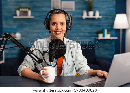 Portrait of smiling blogger looking at camera before starting live video at home studio podcast holding cup of coffee. Content creator blogger woman recording brodcast live streaming for Internet