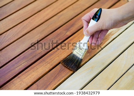 hand holding a brush applying varnish paint on a wooden garden table - painting and caring for wood with oil Royalty-Free Stock Photo #1938432979