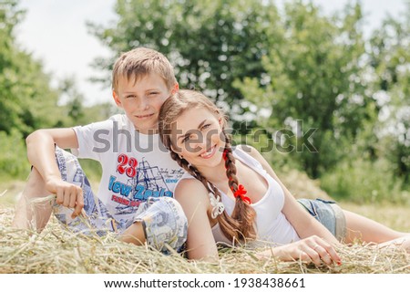 Teenage sister and little brother lying on hay on summer day outdoors, looking at camera and smiling