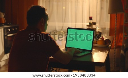 Young man watching video from computer on kitchen table with closed night lights. Young man drinking coffee, watching video from computer with watch point on green screen. 