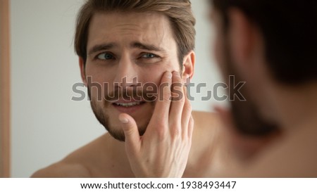 Close up head shot unhappy man looking in mirror, feeling stressed of sensitive skin or acne breakout, thinking of cosmetology treatment. Depressed young guy dissatisfied with skin condition. Royalty-Free Stock Photo #1938493447