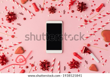 Top view photo of birthday composition with red ribbon stars serpentine confetti bright decorations and smartphone with copyspace in the middle on pastel pink background