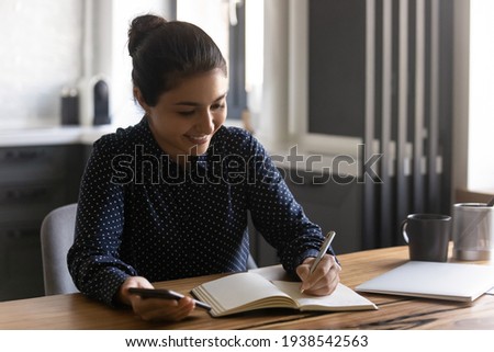 Smiling millennial Indian female student write note in notebook working or studying online on smartphone. Happy young ethnic woman use cellphone make list or plan in paper notepad. Management concept. Royalty-Free Stock Photo #1938542563