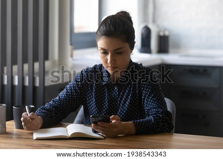 Focused young Indian woman write in notebook study distant at home using smartphone gadget. Smart concentrated ethnic female make notes in notepad, look at cellphone screen planning thinking. Royalty-Free Stock Photo #1938543343