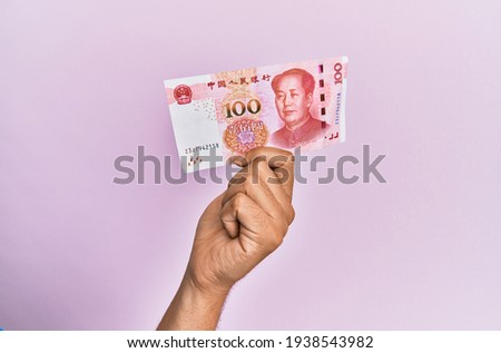 Hispanic hand holding 100 chinese yuan banknote over isolated pink background.