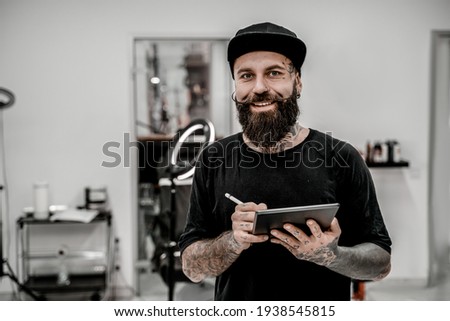 Young male tattoo artist with beard holding pencil and sketch looking positive and happy standing and smiling in workshop place. Royalty-Free Stock Photo #1938545815
