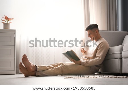 Man with cup of hot drink reading book at home. Floor heating concept Royalty-Free Stock Photo #1938550585