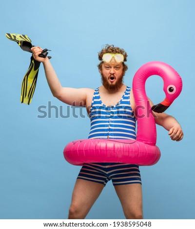Action, dancing. Cute funny red-headed man in swimming suit and swim ring flamingo isolated on blue background. Retro style. Concept of facial expression, holidays, funny meme emotions.