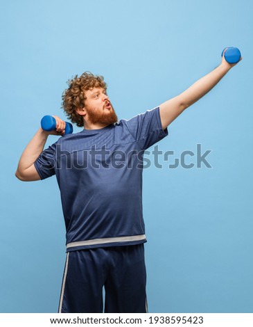 Sport life. Funny red-headed man in blue sports uniform with weights training isolated on blue background. Concept of human facial expression, action, funny meme emotions. Copy space for ad.