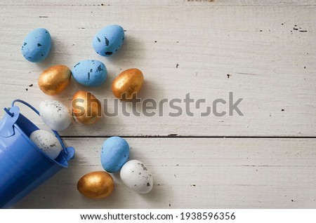 Correct Golden, white and blue Easter eggs with blue bucket on rustic wooden white background. Trendy Easter composition. Greeting card, poster, flyer, promotion, article. Copyspace.  Selective focus.